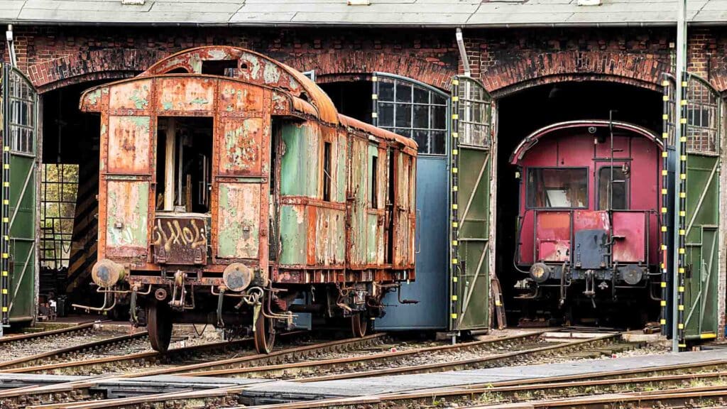 Dois vagões de trem antigos, exibindo ferrugem e desgaste, estão estacionados em trilhos de trem em frente a uma estrutura de arco de tijolos. Ecoando a nostalgia de Tempo de Transporte, eles servem como um lembrete atemporal em meio ao mundo em evolução que se aproxima de 2025.