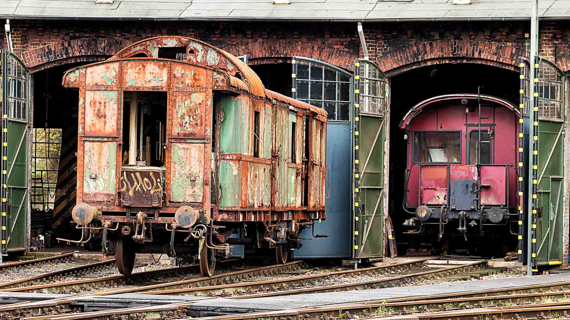 Dois vagões de trem antigos, exibindo ferrugem e desgaste, estão estacionados em trilhos de trem em frente a uma estrutura de arco de tijolos. Ecoando a nostalgia de Tempo de Transporte, eles servem como um lembrete atemporal em meio ao mundo em evolução que se aproxima de 2025.