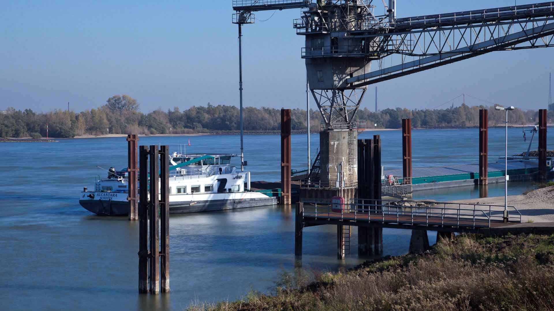 Um barco está atracado em uma instalação de carga industrial perto de um rio, com estruturas de metal e um guindaste. Conforme 2025 se aproxima, este local se adapta às tendências de importação. Árvores e uma ponte são visíveis ao fundo, misturando indústria com a calma da natureza.