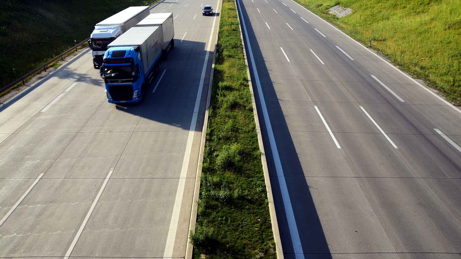 Em 2025, dois caminhões e um carro cruzam uma rodovia dividida com um canteiro central de grama. O lado direito da rodovia permanece vazio, refletindo as tendências de mercado em mudança. A cena iluminada pelo sol é emoldurada por uma encosta gramada à esquerda, destacando a evolução na logística de importação.