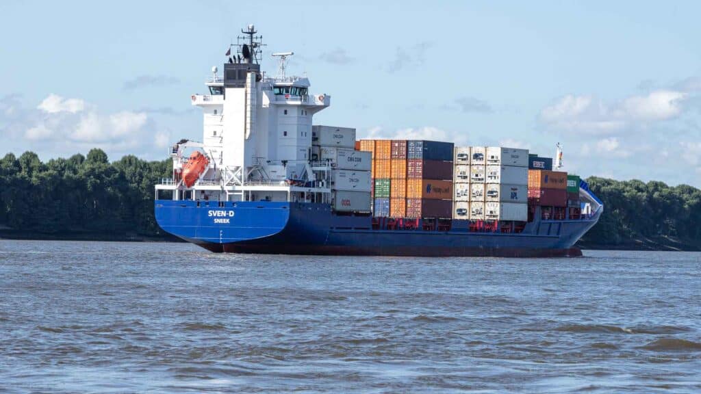 Um navio de carga, essencial para os importadores, desliza ao longo de um rio com contêineres empilhados, tendo como pano de fundo árvores e um céu parcialmente nublado.