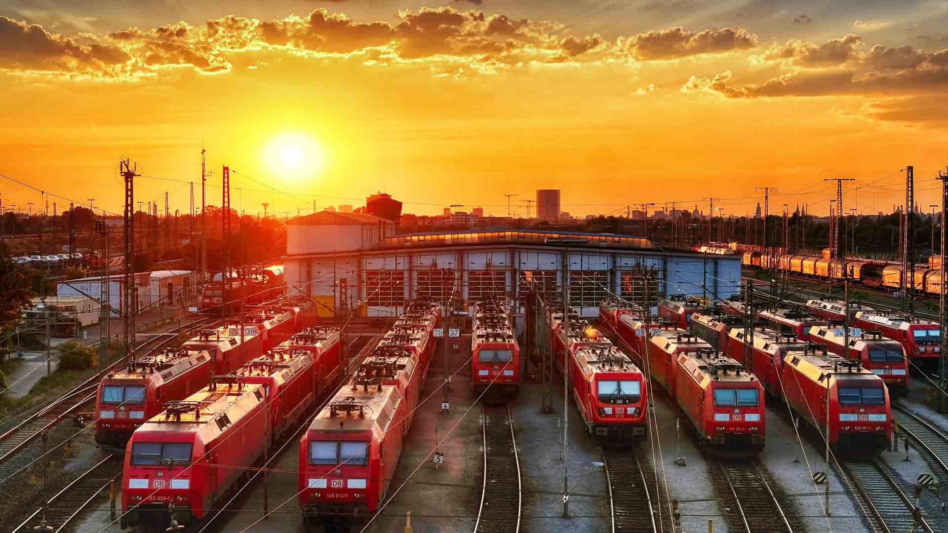 Trens vermelhos estacionados em vários trilhos em frente a um grande galpão ao pôr do sol, com o horizonte da cidade e pilhas de contêineres sugerindo um comércio movimentado ao fundo.