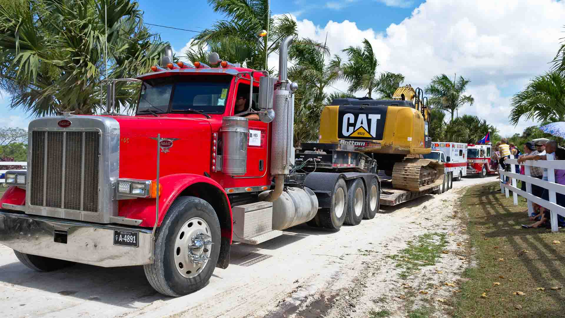 Um caminhão vermelho transporta meticulosamente uma escavadeira CAT amarela em um trailer de plataforma ao longo de uma estrada de terra ladeada por palmeiras, uma cena em que se poderia imaginar a descoberta de oportunidades para comprar uma máquina tão impressionante.