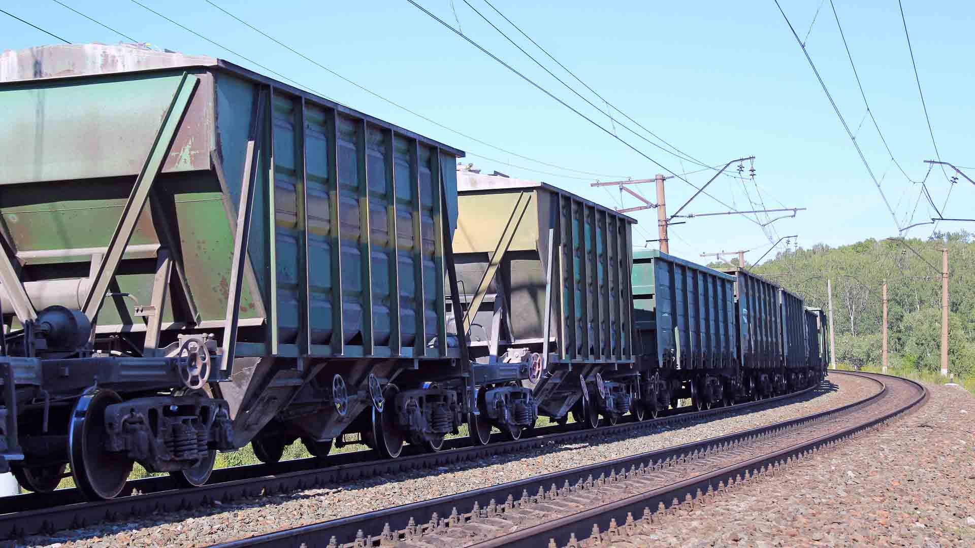 Um longo trem de carga com vagões abertos, transportando bens preciosos talvez para importar da China, viaja em uma linha férrea, cercado por vegetação, sob um céu azul claro. A cena é um testamento do valor máximo da logística moderna em 2024.
