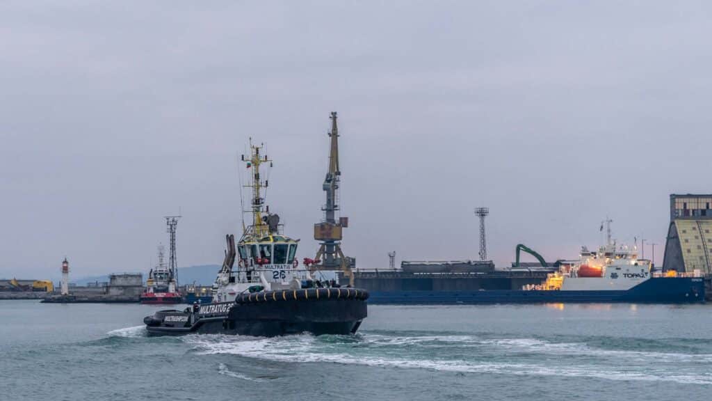 Rebocadores guiam habilmente um navio de carga perto de uma doca, com guindastes e edifícios industriais pairando ao fundo sob um céu nublado. Esta cena simboliza os processos vitais de 2024, onde importar produtos da China se torna essencial para atender às demandas globais.