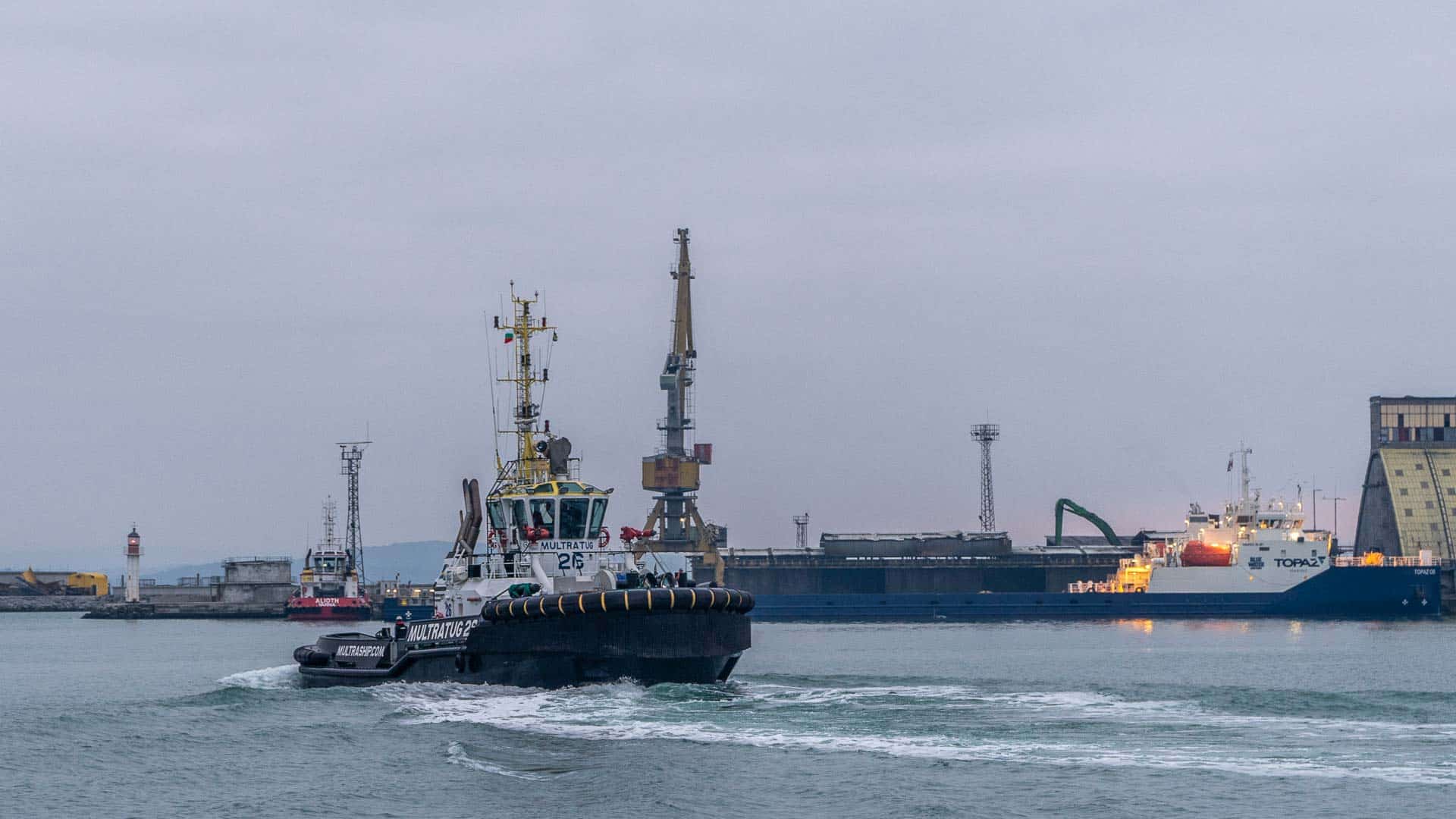 Rebocadores guiam habilmente um navio de carga perto de uma doca, com guindastes e edifícios industriais pairando ao fundo sob um céu nublado. Esta cena simboliza os processos vitais de 2024, onde importar produtos da China se torna essencial para atender às demandas globais.