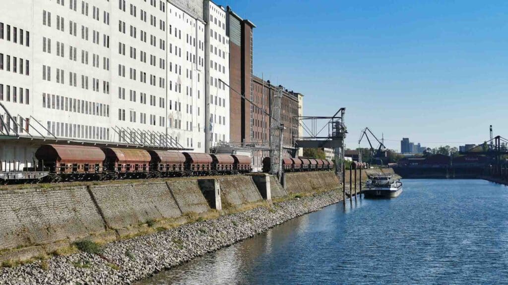 Um trem de carga desliza ao longo dos trilhos ribeirinhos ao lado de edifícios industriais; um barco navega no rio enquanto guindastes, essenciais nos processos de importação, aparecem ao fundo sob um céu azul claro.