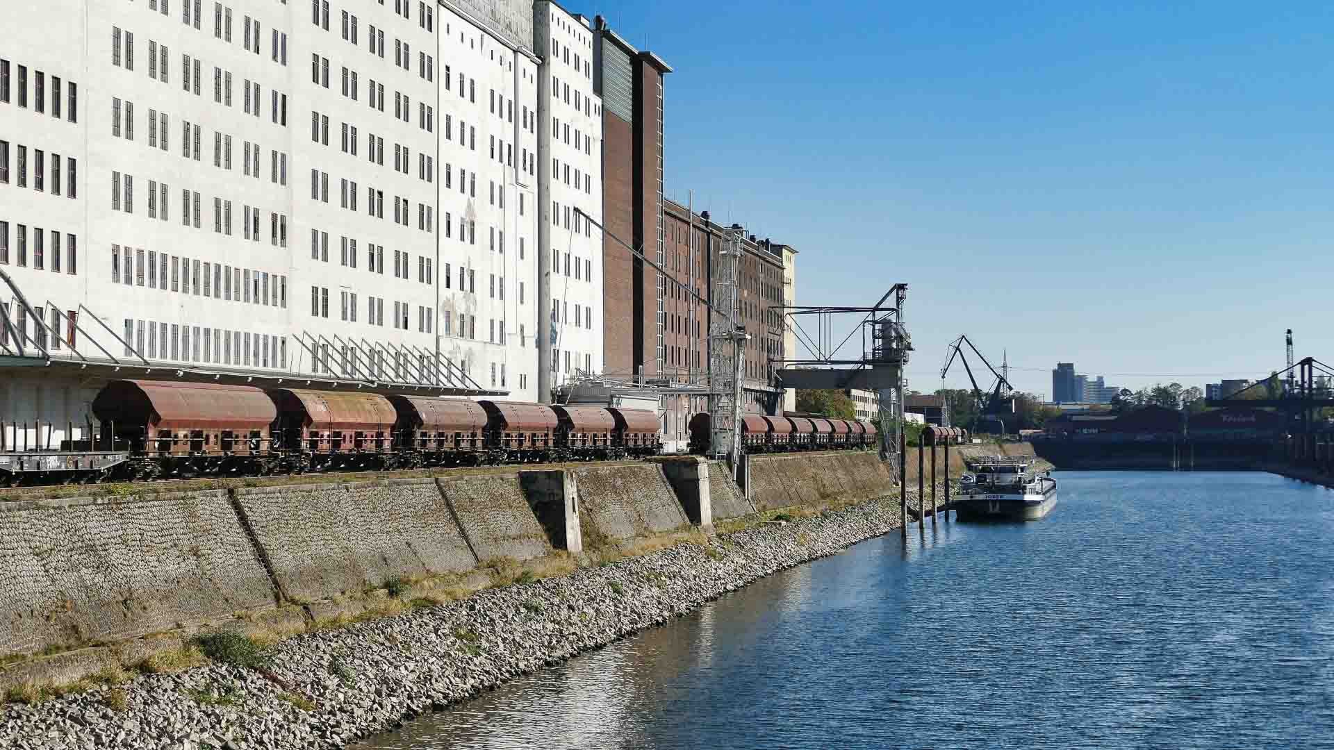 Um trem de carga desliza ao longo dos trilhos ribeirinhos ao lado de edifícios industriais; um barco navega no rio enquanto guindastes, essenciais nos processos de importação, aparecem ao fundo sob um céu azul claro.