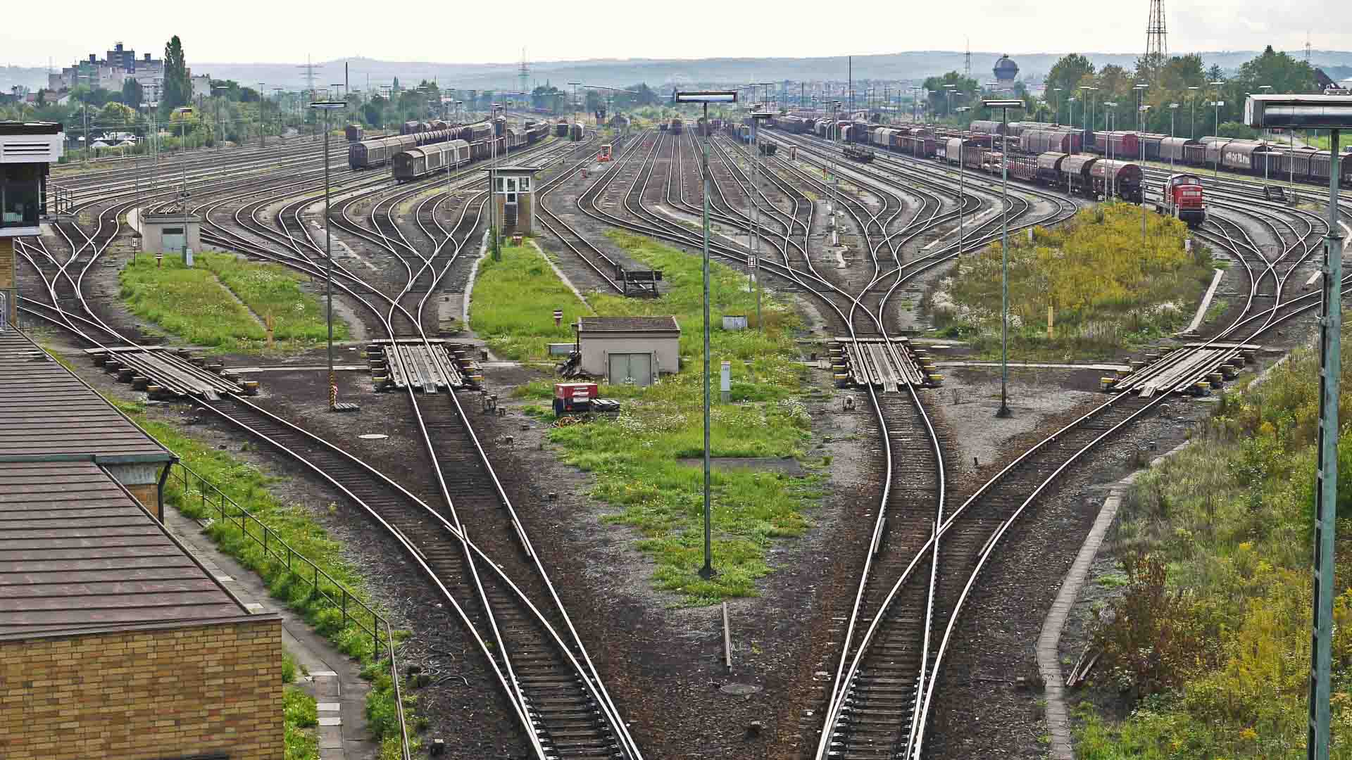 Vista aérea de um complexo pátio ferroviário com vários trilhos de trem divergindo e convergentes, cercado por grama verde e trens de carga, movimentados com a importação da China.