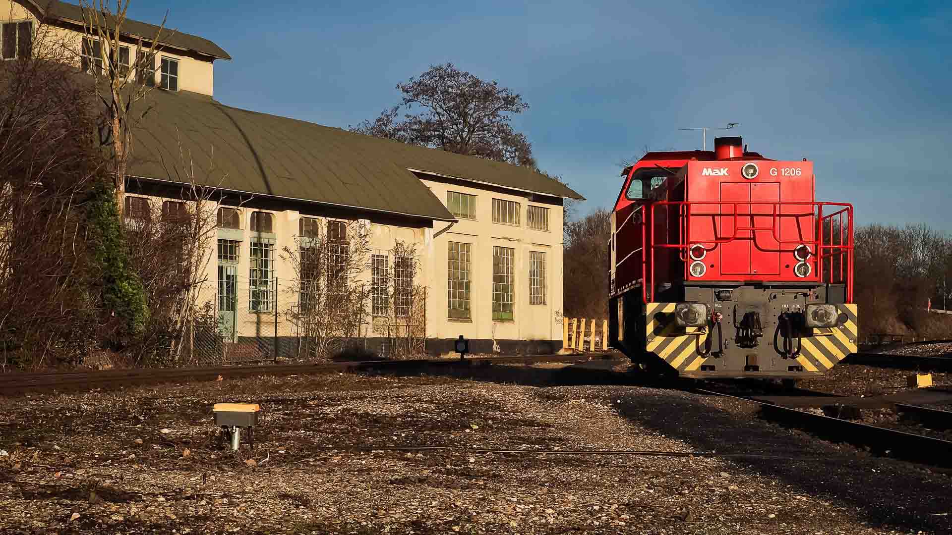 Uma locomotiva vermelha está nos trilhos de uma ferrovia ao lado de um prédio industrial sob um céu parcialmente nublado, simbolizando os benefícios da importação para negócios com sua integração perfeita ao comércio global.