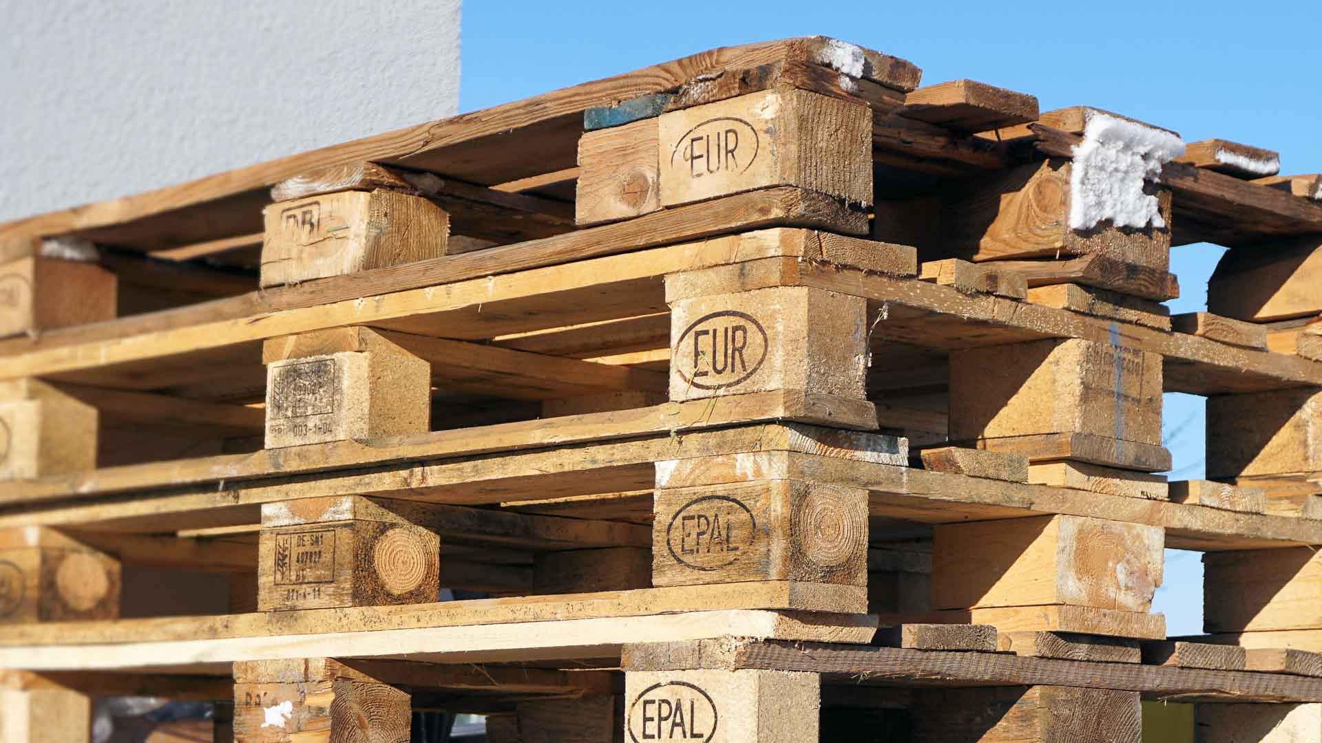 Uma pilha de paletes de madeira, cada uma marcada com os carimbos "EUR" e "EPAL", ergue-se orgulhosamente contra o céu azul claro, pronta para importar mercadorias com eficiência em 2024, mantendo os custos baixos.