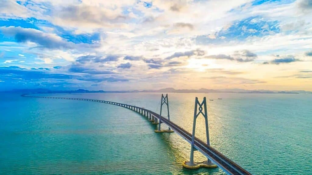 Uma longa ponte se estende sobre um vasto corpo de água, sob um céu parcialmente nublado com um pôr do sol ao fundo, lembrando uma lista de verificação de importação para garantir que tudo esteja pronto para a jornada que temos pela frente.