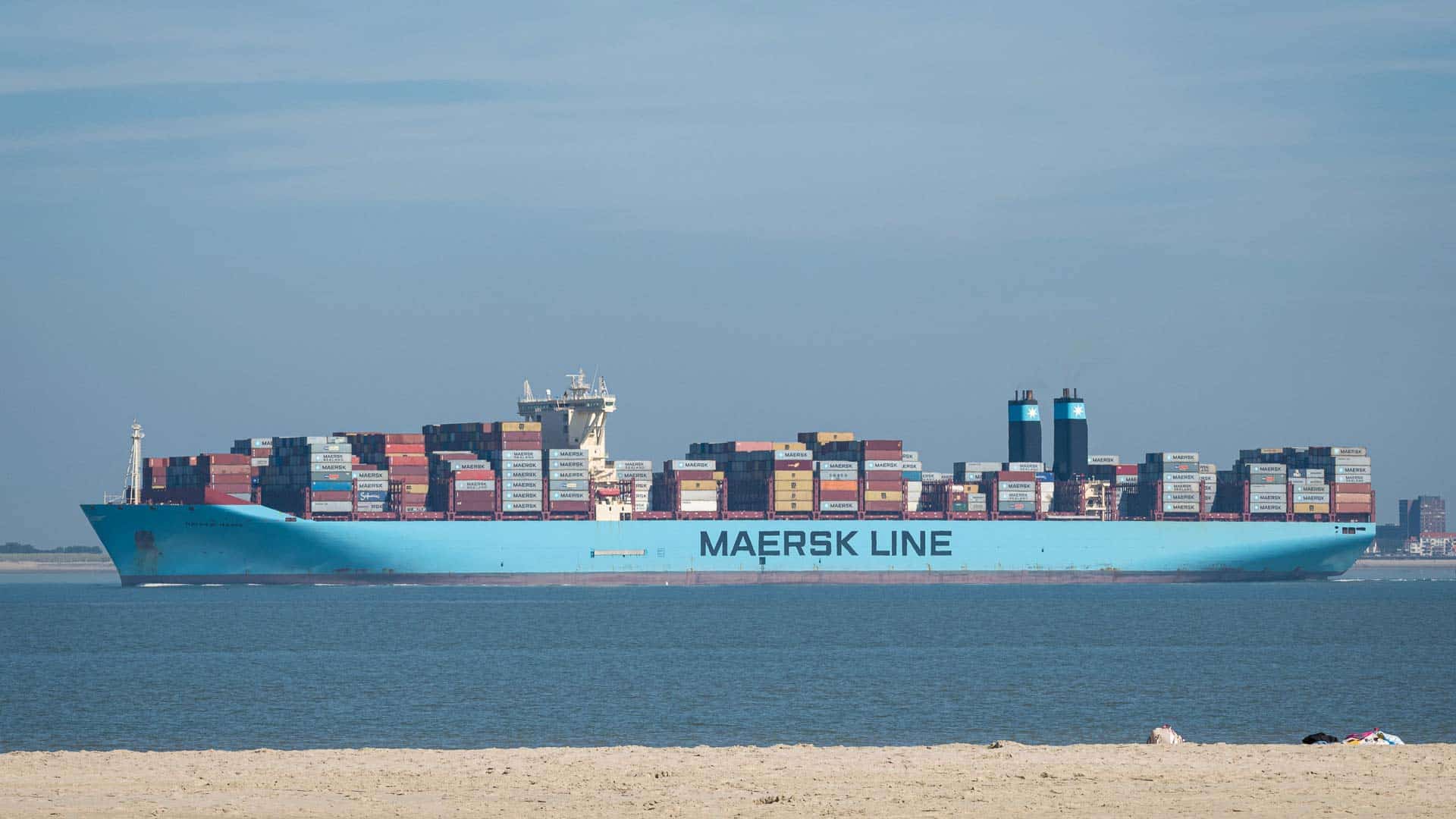 Um grande navio porta-contêineres azul com a etiqueta "Maersk Line" navega em águas calmas perto de uma costa arenosa sob um céu limpo, seu casco carregado com mercadorias para importação, cada item meticulosamente organizado para uma classificação fiscal precisa.