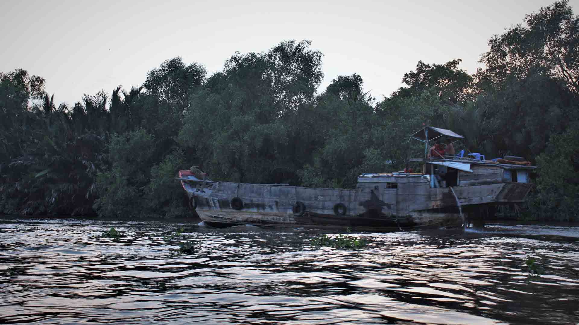 Um barco rústico está atracado na margem de um rio com uma vegetação verdejante ao fundo, onde os efeitos da Taxa Selic sutilmente ondulam através das fronteiras como esses reflexos serenos na água sob um céu nublado.
