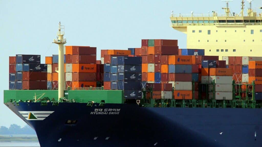 Visão de perto do grande navio porta-contêineres "Hyundai Drive", exibindo um movimentado negócio com vários contêineres empilhados em seu convés, prontos para navegar pelas mudanças no mercado.