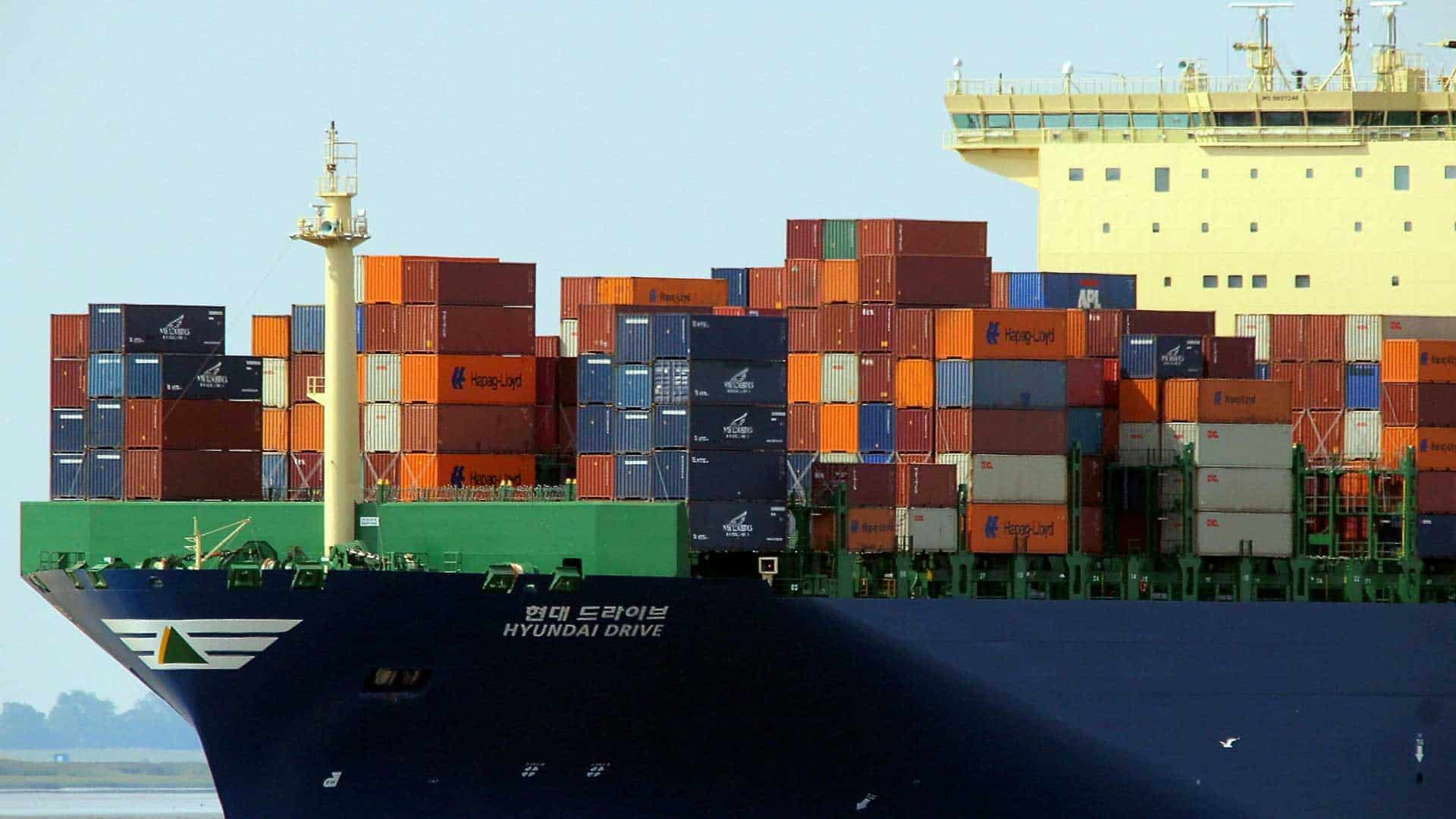 Visão de perto do grande navio porta-contêineres "Hyundai Drive", exibindo um movimentado negócio com vários contêineres empilhados em seu convés, prontos para navegar pelas mudanças no mercado.