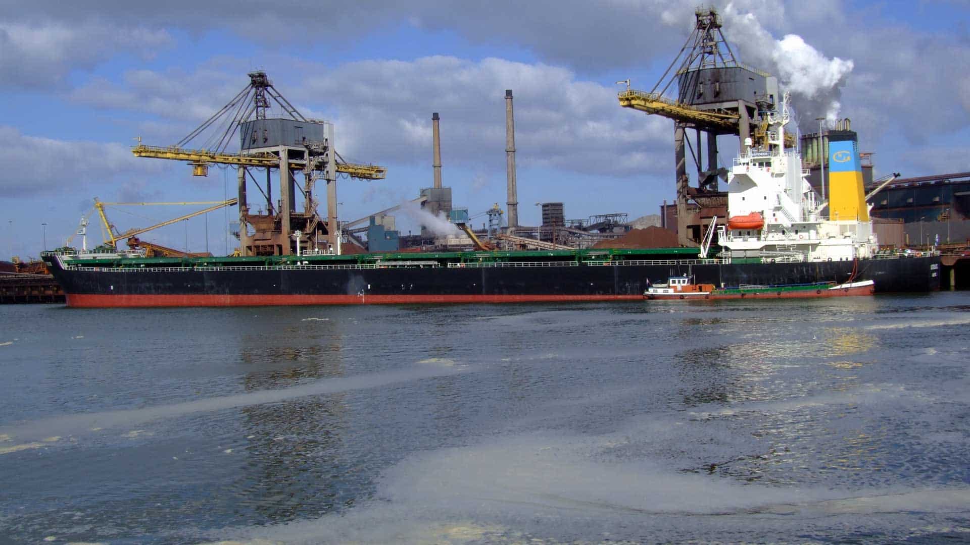 Um grande navio de carga carregado com produtos importados atracou em um porto industrial com guindastes imponentes, ao lado de um rebocador menor em um dia nublado.