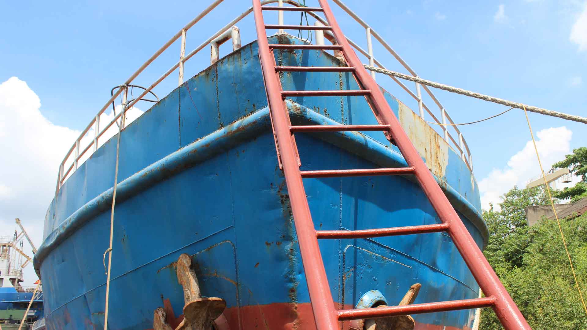 Um grande navio azul, essencial para a importação, está atracado e visto da proa. Uma escada vermelha se apoia nele, estendendo-se até o convés. O céu limpo com algumas nuvens sugere um dia preparado para aumentar a eficiência nas operações marítimas.