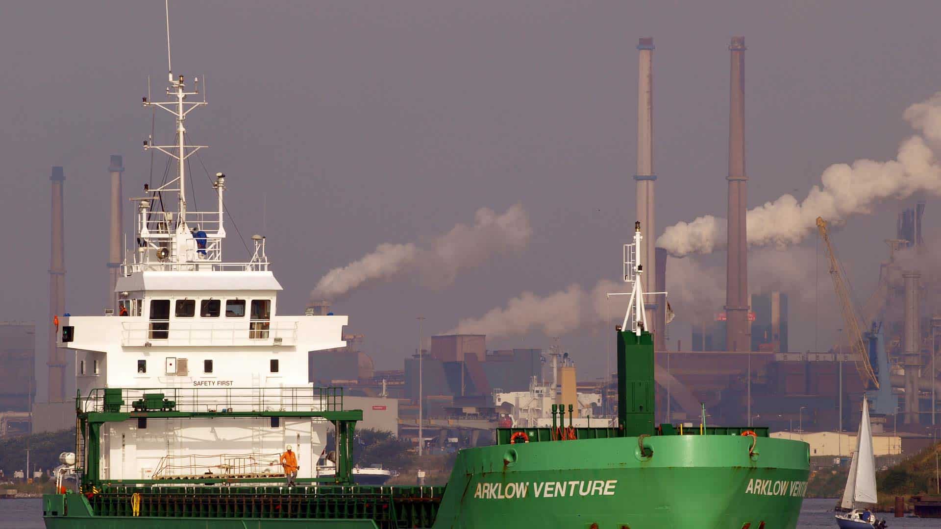 O navio de carga verde "Arklow Venture" desliza sobre a água, seu casco refletindo o poder industrial. Chaminés de fumaça se erguem ao fundo, sugerindo uma importação movimentada que visa aumentar a margem de lucro neste cenário marítimo próspero.
