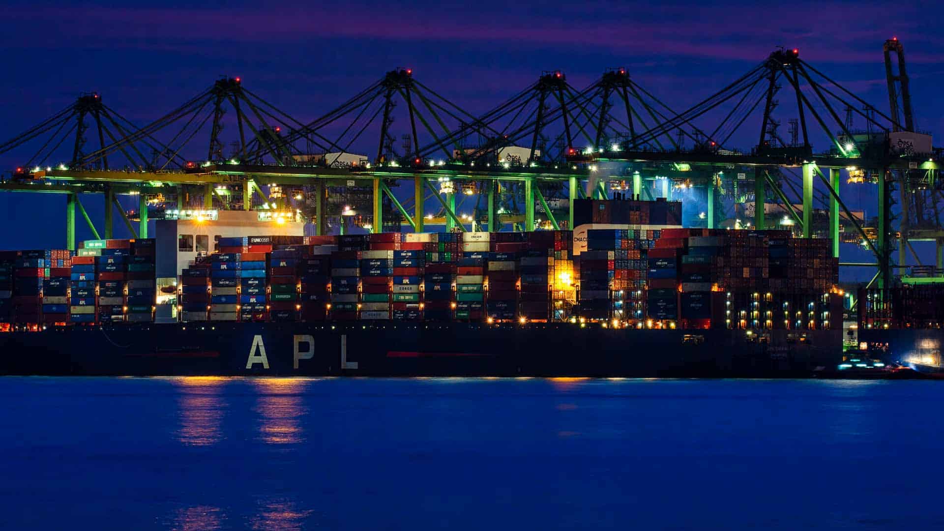 Um navio de carga com contêineres empilhados, fornecidos por fornecedores chineses, está atracado em um porto sob luzes brilhantes, enquanto guindastes pairam no alto durante o anoitecer.
