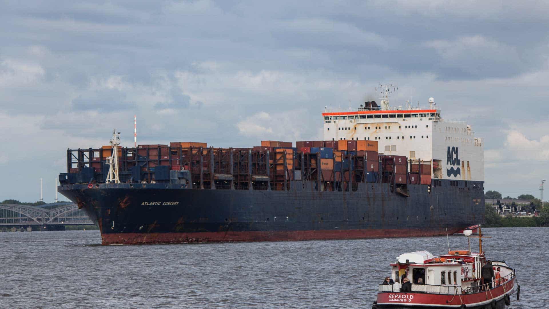 Um grande navio porta-contêineres navega em um rio, com um barco menor próximo em primeiro plano. Enquanto o céu nublado se aproxima, aqueles que buscam "Como Encontrar" fornecedores garantidos podem descobrir que 2024 será seu ano de descoberta em meio a essas vastas remessas.