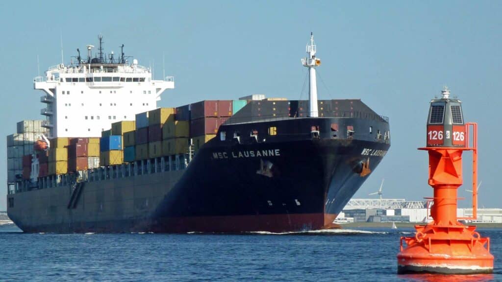 O grande navio de carga, MSC Lausanne, está navegando por um corpo de água, pesadamente carregado com contêineres coloridos cruciais para importar. Uma bóia de navegação vermelha se destaca no primeiro plano.