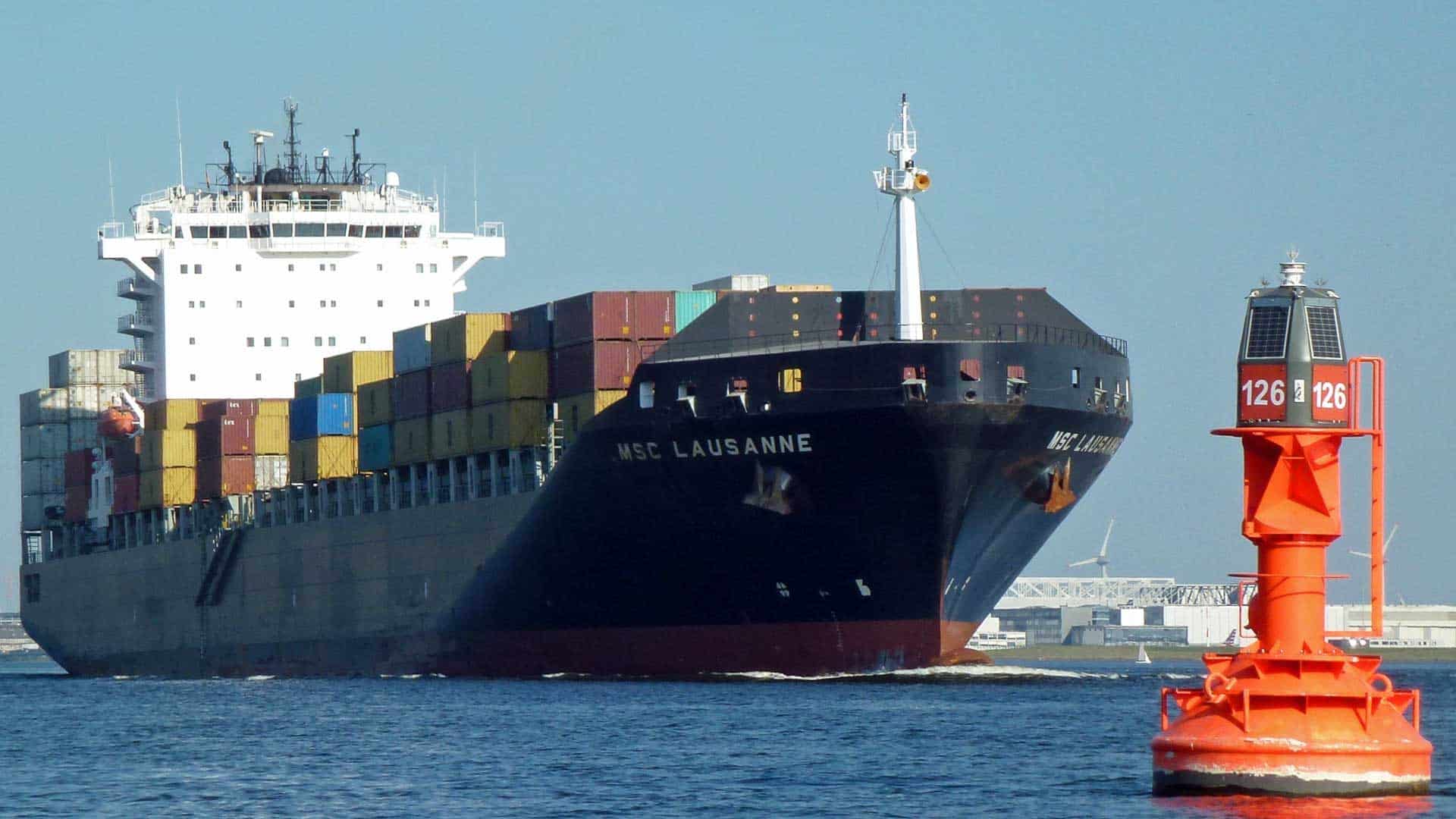 O grande navio de carga, MSC Lausanne, está navegando por um corpo de água, pesadamente carregado com contêineres coloridos cruciais para importar. Uma bóia de navegação vermelha se destaca no primeiro plano.