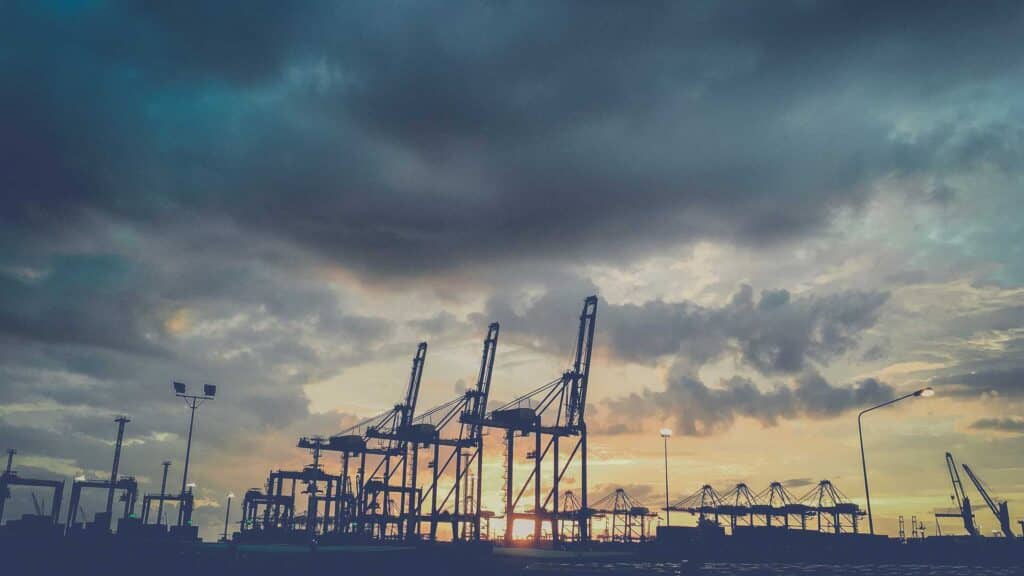 Em 2025, guindastes em uma silhueta portuária contra um céu dramático de pôr do sol com nuvens, incorporando as complexidades da importação e destacando a importância de como evitar riscos em um ambiente comercial movimentado.