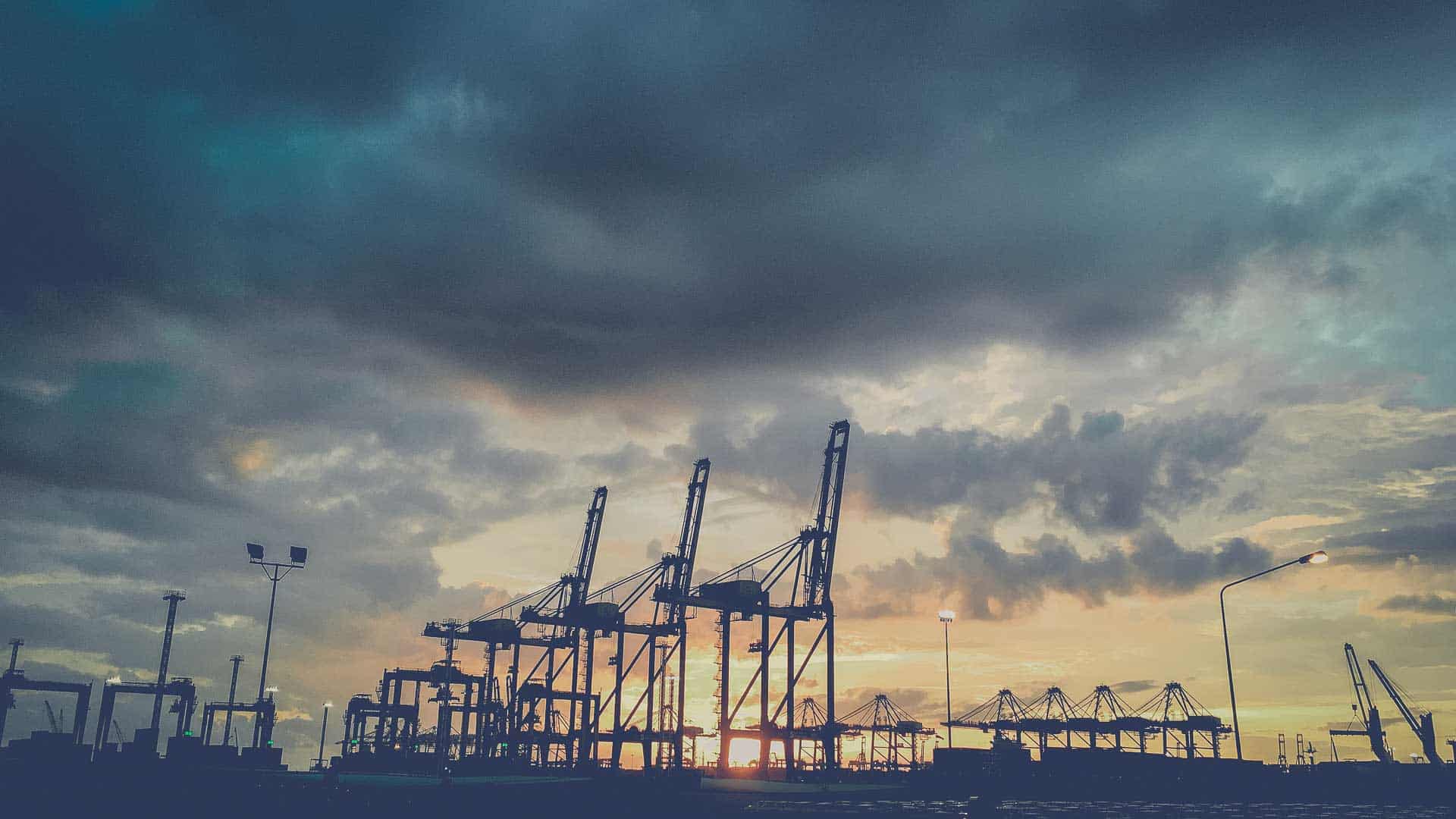 Em 2025, guindastes em uma silhueta portuária contra um céu dramático de pôr do sol com nuvens, incorporando as complexidades da importação e destacando a importância de como evitar riscos em um ambiente comercial movimentado.