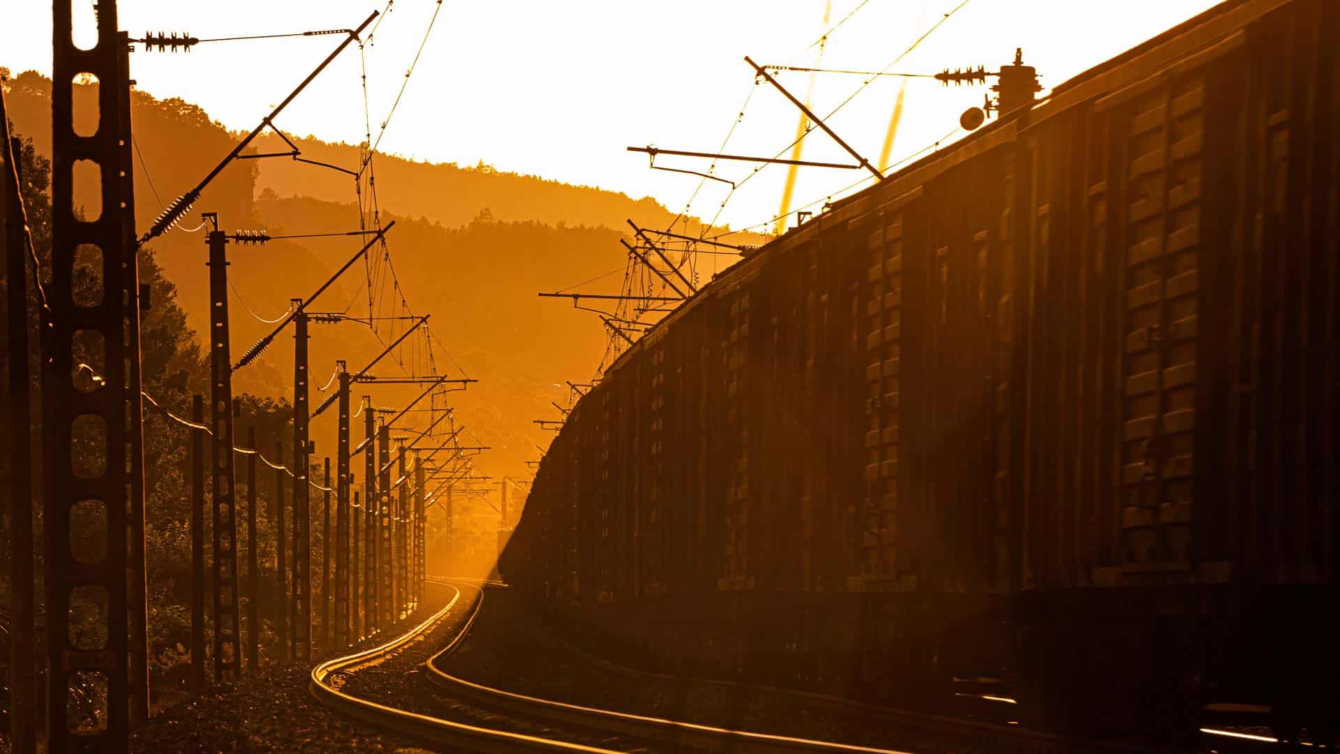 Um trem de carga viaja sobre trilhos ferroviários ao pôr do sol, garantindo qualidade com linhas de energia aéreas e uma paisagem montanhosa ao fundo.