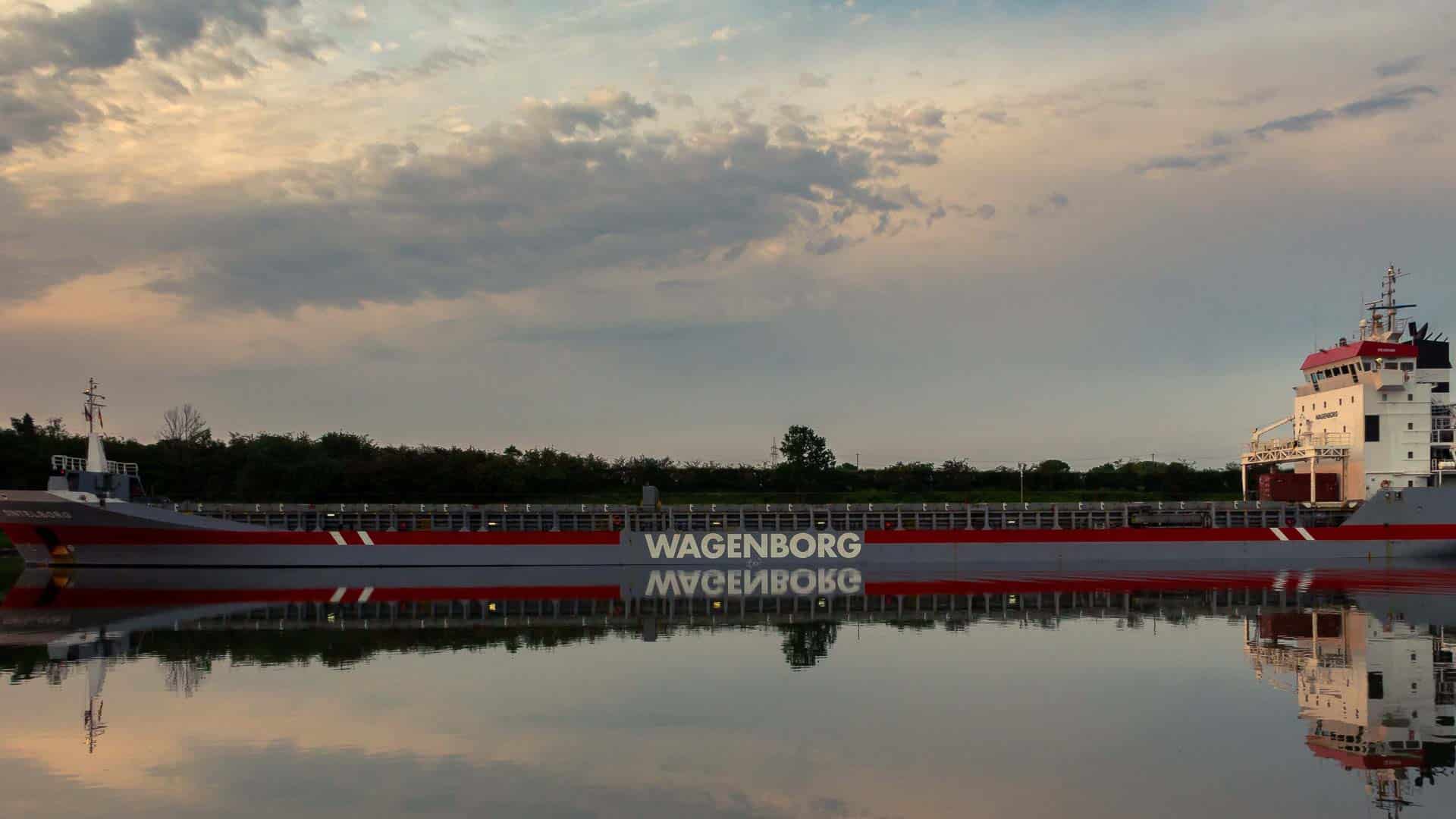 Um navio de carga com "Wagenborg" visível na lateral reflete em águas calmas sob um céu nublado, simbolizando a promessa de importar mercadorias de forma eficiente e com custo reduzido até 2025.