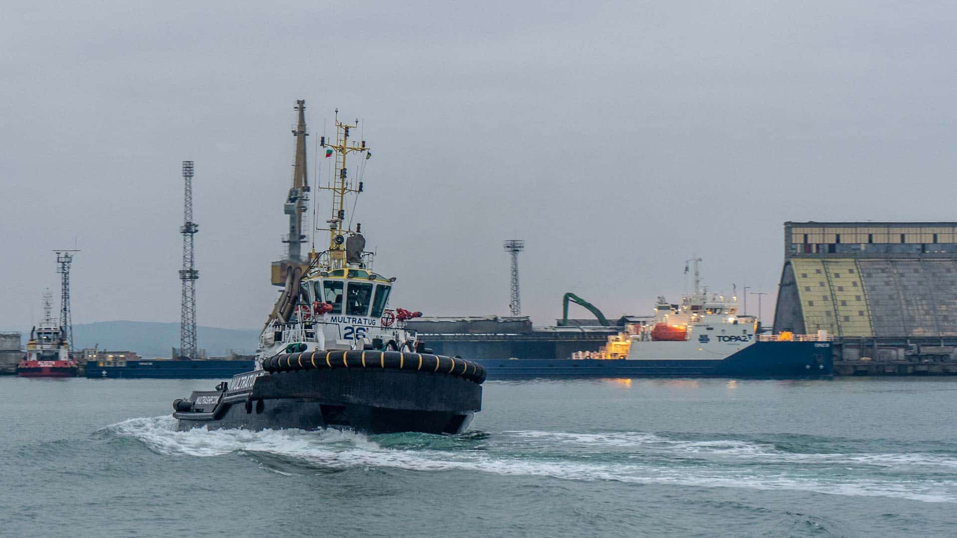 Um rebocador navega por um porto, guiando navios industriais em meio a guindastes imponentes em um dia nublado, como se seguisse um Guia Passo a Passo invisível para garantir operações de importação perfeitas.