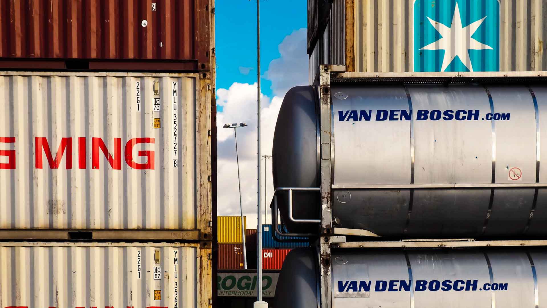 Contêineres de transporte, simbolizando estratégias práticas para gerenciar custos em meio a um ambiente de dólar alto, são empilhados próximos uns dos outros. Esses contêineres apresentam diferentes nomes de empresas e designs vibrantes contra um fundo de céu claro, destacando as complexidades de escolher importar com eficiência.