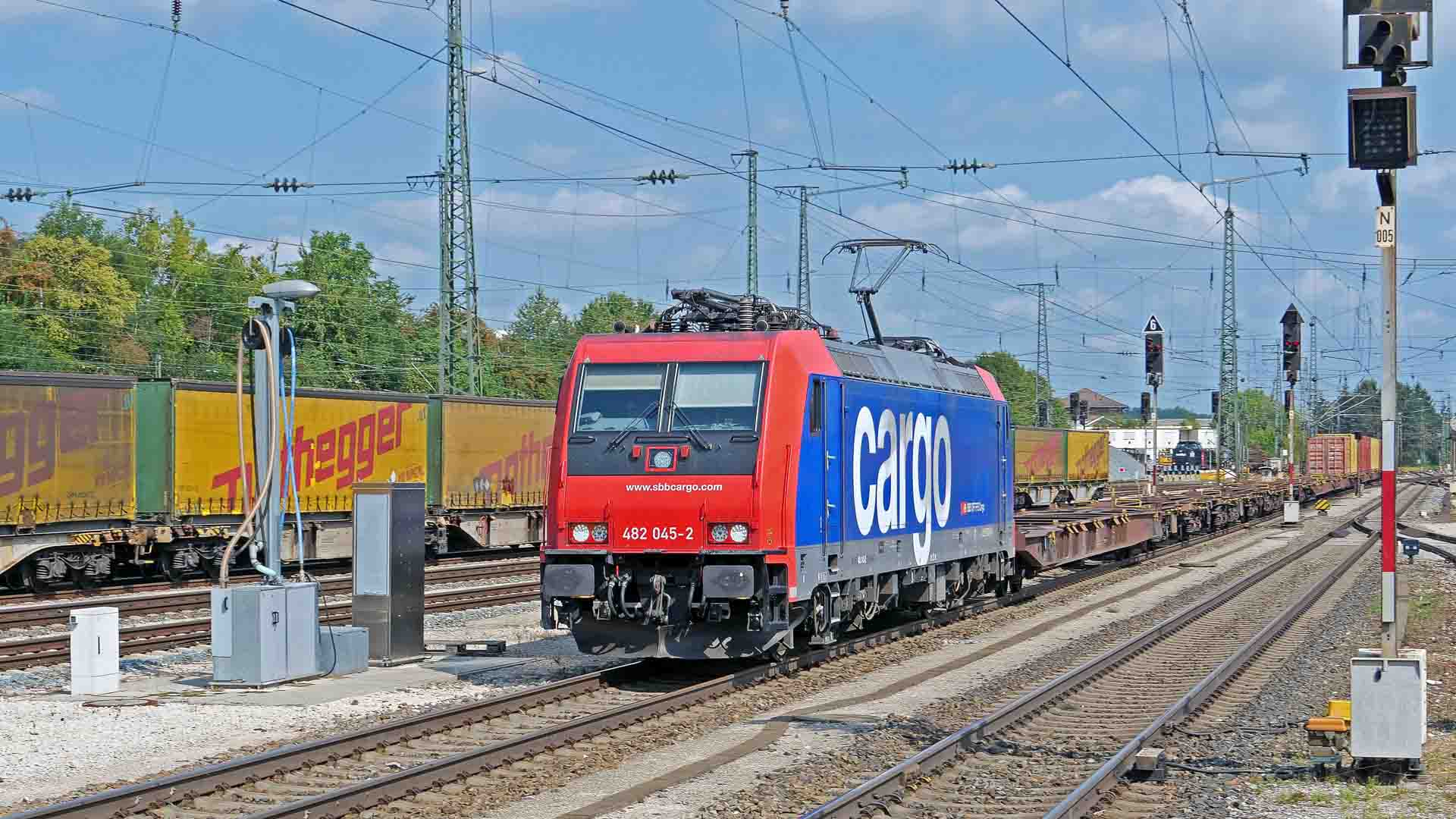 Um trem de carga sobre trilhos com linhas de energia aéreas desliza entre vagões de contêineres e árvores sob um céu parcialmente nublado, exibindo perfeitamente a arte da importação, trazendo mercadorias sem esforço de lugares como a China.