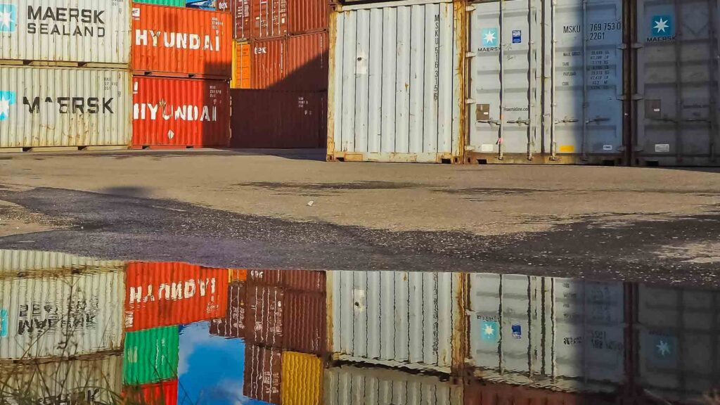 Contêineres de transporte empilhados em uma área portuária, refletidos em uma grande poça no chão, destacam a importância da segurança ao importar do Alibaba.
