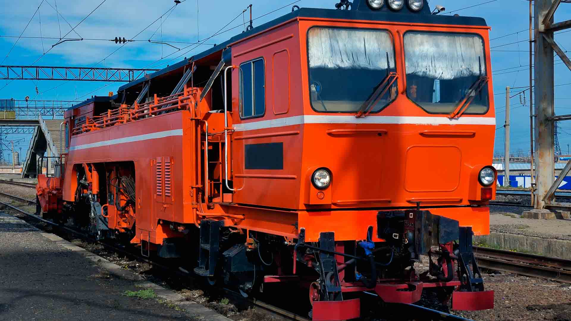 Veículo de manutenção ferroviária laranja brilhante em trilhos de trem, com equipamentos laterais e luzes dianteiras, estacionado perto de uma plataforma ferroviária com cabos aéreos visíveis — um recurso vital, como importar produtos especializados para revenda.