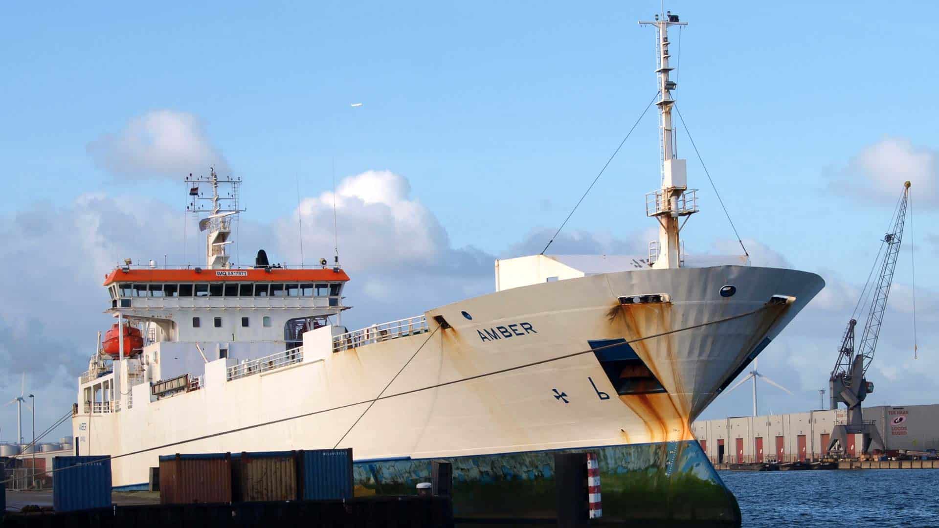 Um navio de carga, que lembra o movimentado comércio do AliExpress, está atracado no porto com seu casco levemente enferrujado. Um guindaste e contêineres de transporte estão prontos ao fundo, silhuetados contra um céu nublado enquanto se preparam para importar legalmente em 2024.