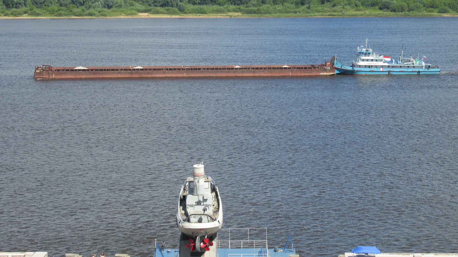 Um rebocador azul empurra uma longa barcaça em um rio largo, lembrando as rotas movimentadas onde as mercadorias são importadas legalmente. Um navio desativado é exibido em terra firme no primeiro plano, com uma linha de árvores verde na margem oposta.