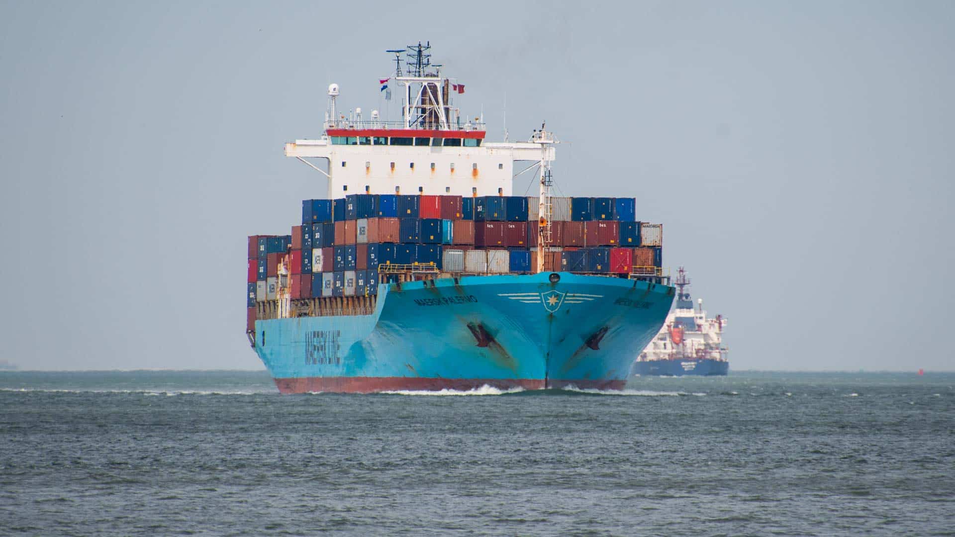 Um grande navio porta-contêineres no oceano, carregado com contêineres coloridos destinados a importar móveis. Outro navio é visível ao fundo, navegando firmemente enquanto ambos navegam por rotas comerciais globais cruciais.