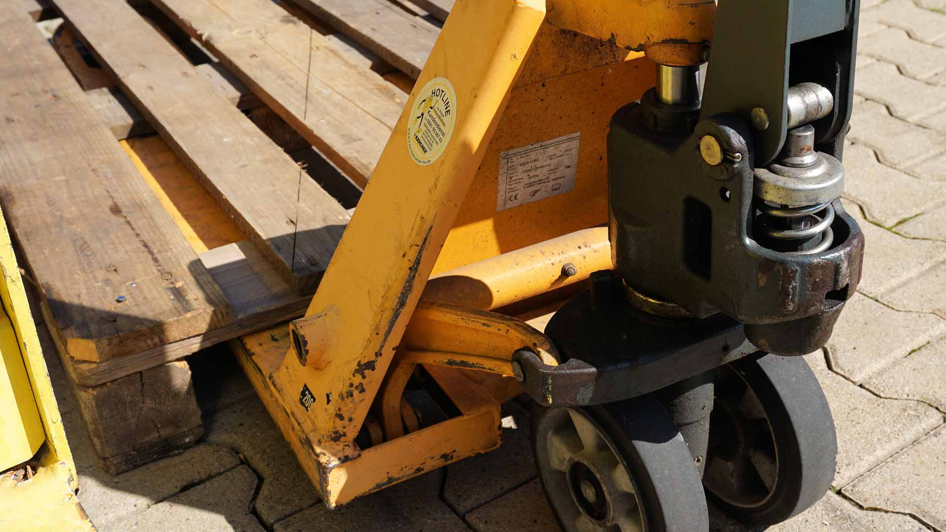 Close-up de um transpaleteiro manual com um palete de madeira sobre uma superfície de tijolos. O transpaleteiro é amarelo com desgaste visível e rodas pretas, pronto para importar pequenas quantidades em 2024.