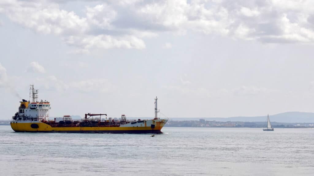 Um grande navio de carga, provavelmente carregado com produtos a importar, navega ao lado de um pequeno veleiro em águas calmas sob um céu nublado, tudo seguindo os padrões de Certificação Inmetro.