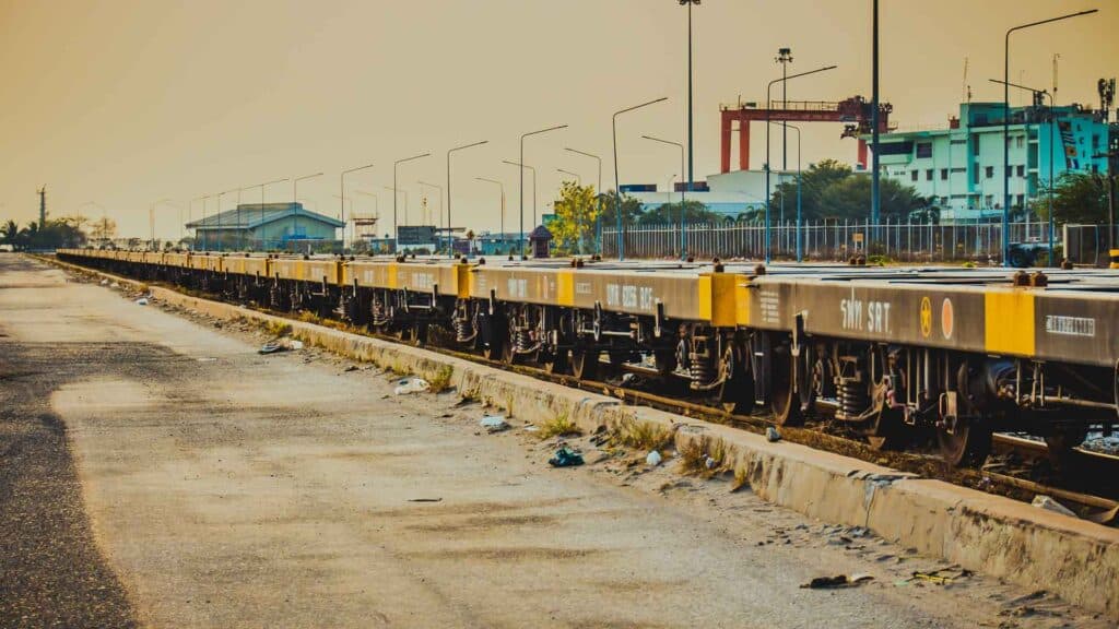 Um longo trem de carga com vagões plataforma, marcado pelo Inmetro, está parado sobre trilhos em uma área industrial, cercado por prédios e guindastes utilizados para transportar produtos.