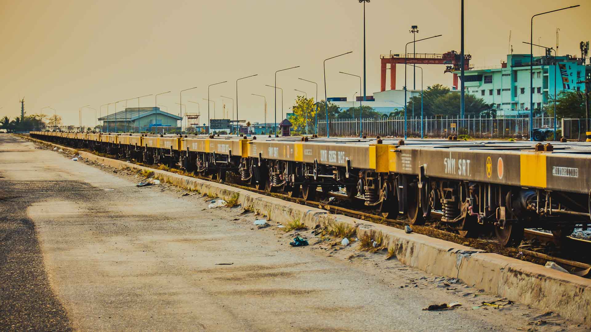Um longo trem de carga com vagões plataforma, marcado pelo Inmetro, está parado sobre trilhos em uma área industrial, cercado por prédios e guindastes utilizados para transportar produtos.