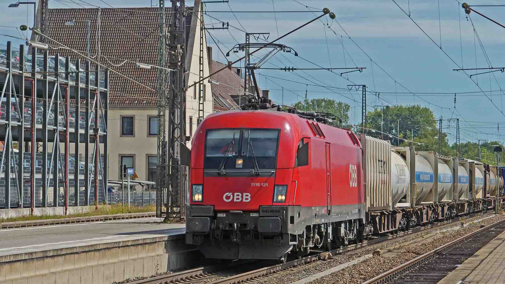 Um trem de carga vermelho ÖBB viaja sobre trilhos ferroviários, aumentando a eficiência da imigração ao passar por uma estação com carros estacionados à esquerda.