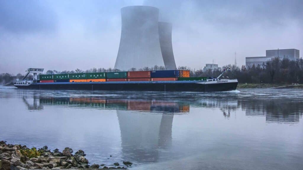Um navio de carga carregado com contêineres viaja em um rio, com duas grandes torres de resfriamento ao fundo refletindo na água. Isso ilustra as movimentadas rotas comerciais usadas para importar da China, exibindo o comércio global e a logística que devem florescer em 2024.