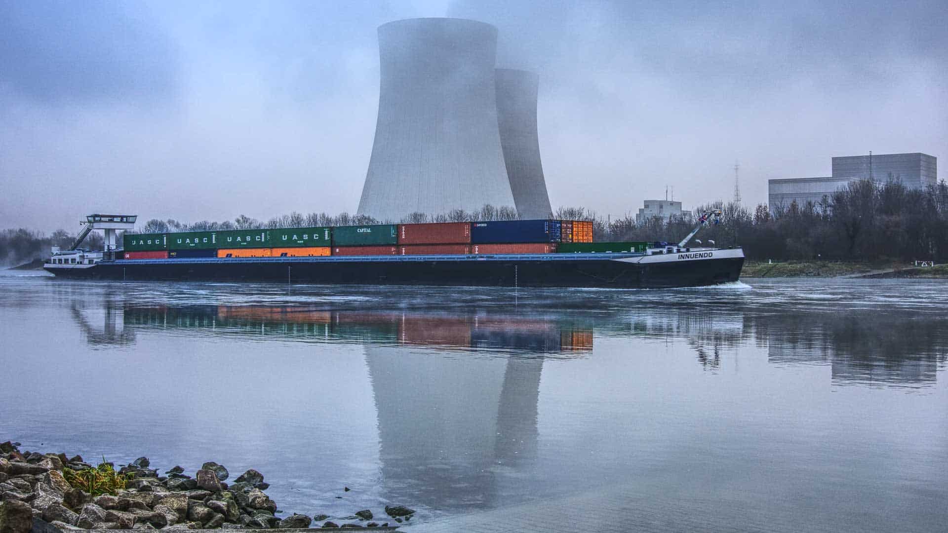 Um navio de carga carregado com contêineres viaja em um rio, com duas grandes torres de resfriamento ao fundo refletindo na água. Isso ilustra as movimentadas rotas comerciais usadas para importar da China, exibindo o comércio global e a logística que devem florescer em 2024.